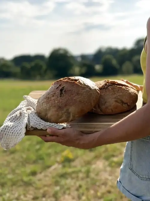Miguela trägt zwei frisch gebackene Sauerteigbrote auf einem Holzbrett über die Wiese